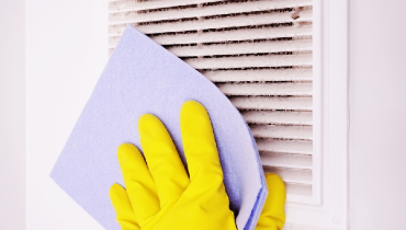 Person wearing yellow rubber gloves using a purple towel to clean a dirty air vent.