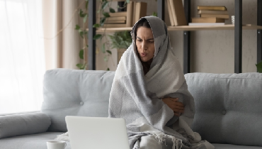 Young woman wrapped in a blanket sitting on her couch into front of a laptop computer.