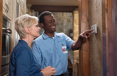 Male Aire Serv service professional adjusting thermostat beside female customer in blue shirt.