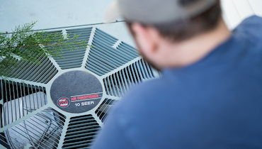 Man in baseball cap and blue t-shirt staring down at outdoor central AC unit condenser fan.