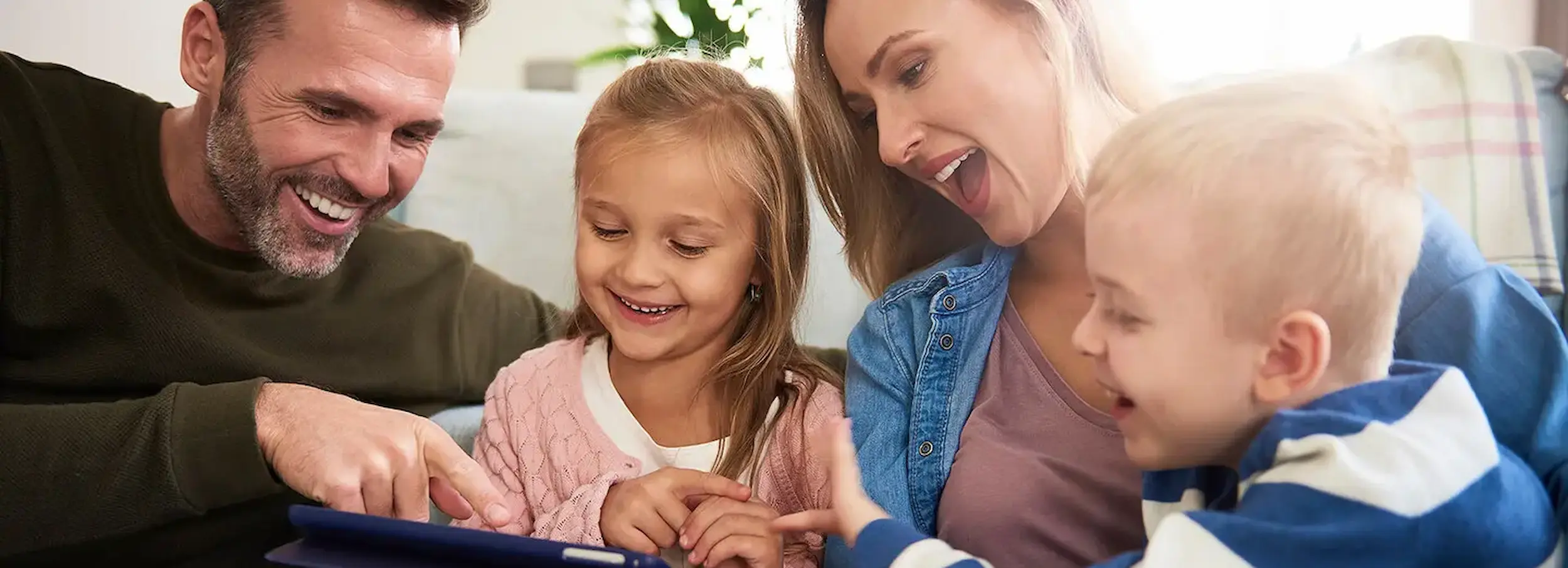 Smiling couple and two children pointing at tablet device in brightly lit living room interior.