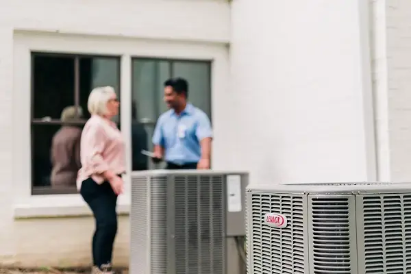 Close up of AC with Aire Serv tech discussing emergency HVAC with client in the background