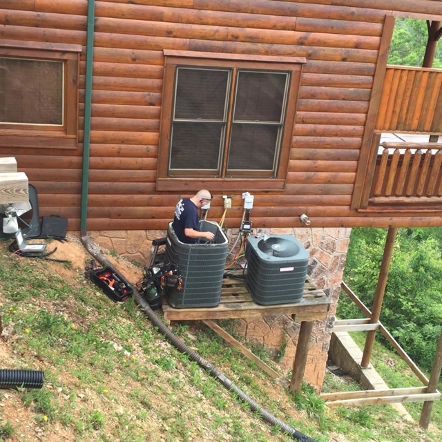 HVAC technician standing on a deck working on an outdoor AC unit at a log cabin