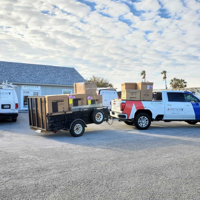 Truck and open trailer loaded with large boxes containing HVAC equipment for installation or service by Aire Serv of Coastal Bend.