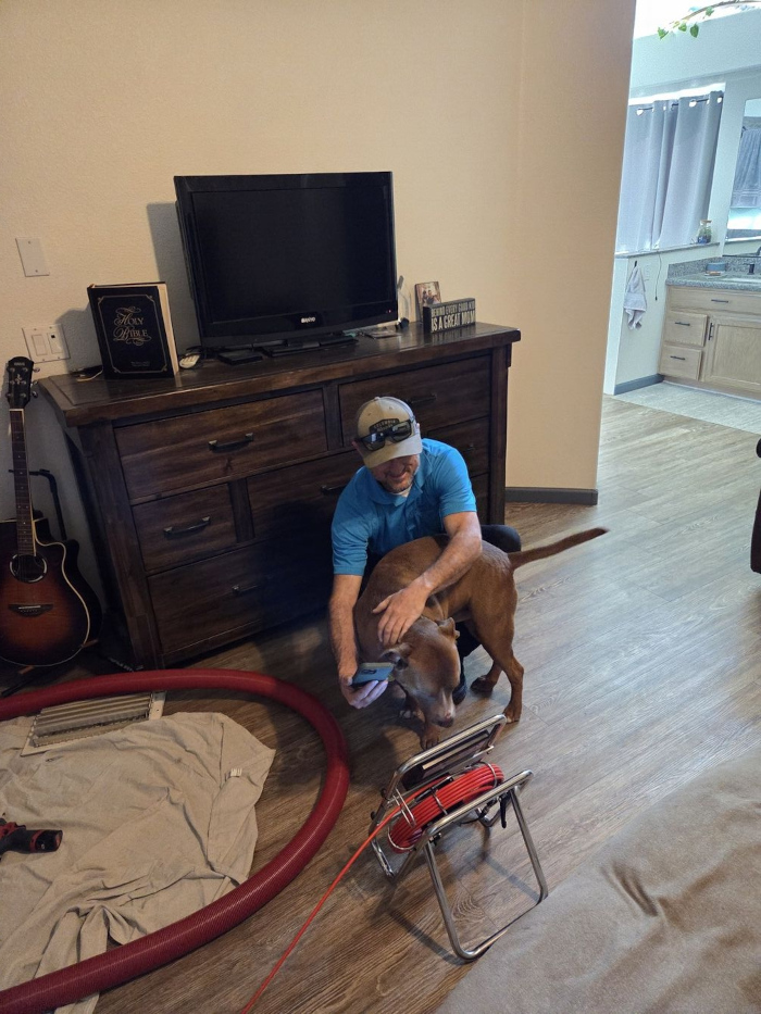 Aire Serv team member Lyle sitting on the floor, reviewing a video feed on a tablet while friendly interacting with a customer's dog.
