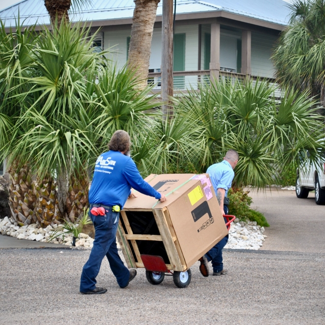 Two Aire Serv of Coastal Bend HVAC technicians moving a large HVAC equipment box outdoors.