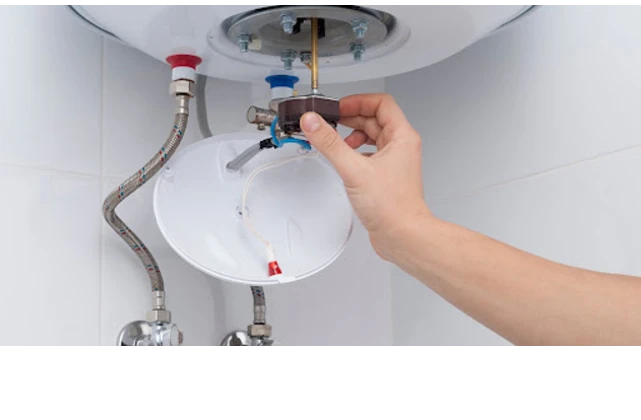 A hand holding the adjustable thermostat knob of a residential water heater with both cold and hot water connections.