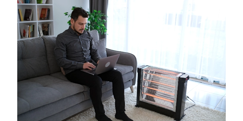 A man works on a laptop next to a portable electric heater.