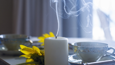 White candle with smoke coming from top sitting next to a tea cup and yellow flowers