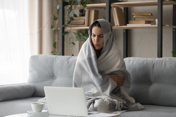 Young woman wrapped in a blanket sitting on her couch into front of a laptop computer.