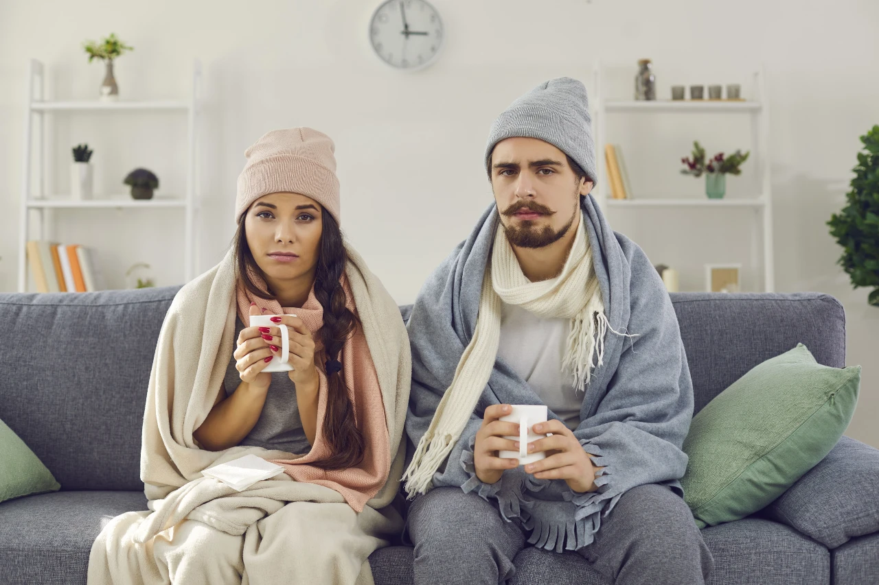 Upset young couple sitting on the couch, bundled up and having problem with central heating