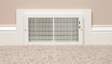 An air duct vent at the bottom of a beige wall next to beige carpet