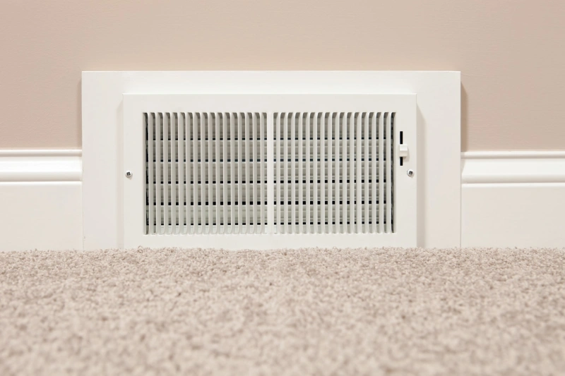 An air duct vent at the bottom of a beige wall next to beige carpet