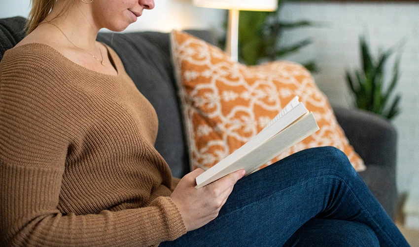 Woman reading on a gray couch next to an orange and white throw pillow with two plants in the background.