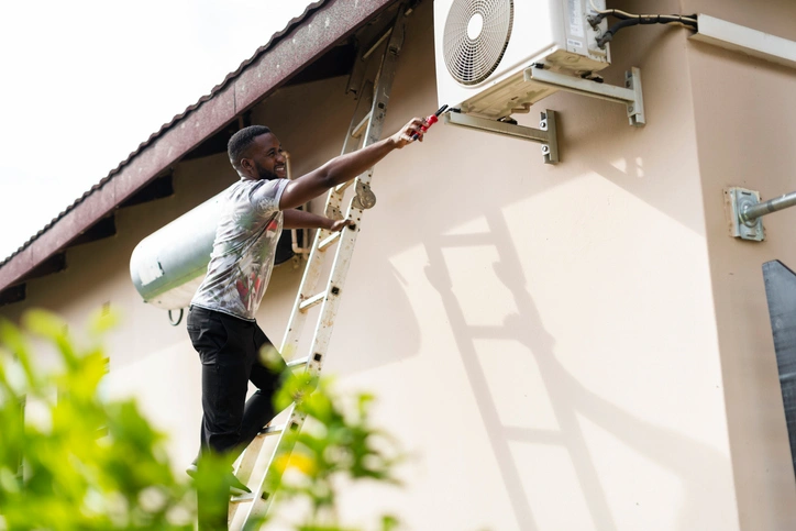 Man standing on ladder outside home and reaching over to the wall-mounted HVAC unit with a screwdriver.