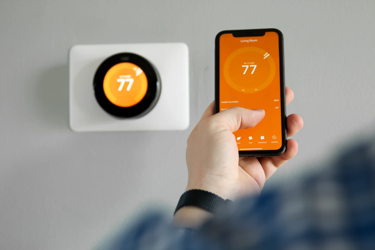 A homeowner controls a thermostat with an app on their cell phone. The image shows both the cell phone screen and the smart thermostat synched at 77 degrees. They are also both sharing an orange background to highlight their connectivity.