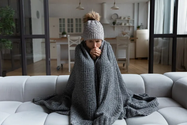 A woman wrapped in a blanket on her couch because her home’s heater is broken.