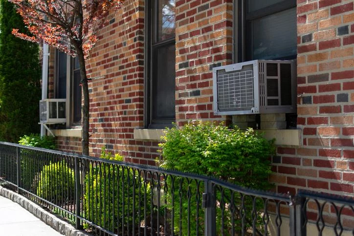 Outdoor window air conditioning units on an old New York City brick apartment building with green plants along a sidewalk | Aire Serv of Springfield, Illinois