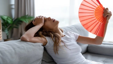 A woman sitting on a couch uses a paper fan to cool herself off during a summer heat wave. | Aire Serv of North Central Arizona