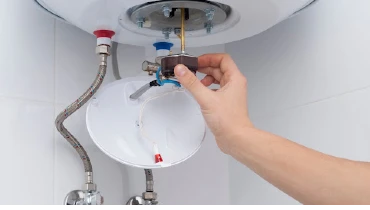A hand holding the adjustable thermostat knob of a residential water heater with both cold and hot water connections.