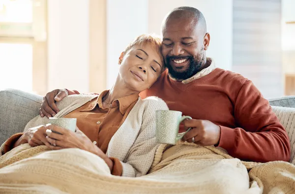 African American couple holding mugs and resting under blanket on living room couch.