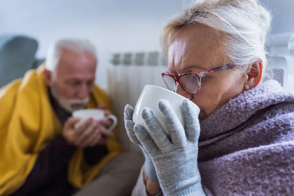 Elderly couple wearing gloves and scarf indoors and drinking from large white mugs.
