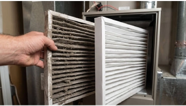 A hand holds a dirty furnace air filter beside a clean replacement filter.