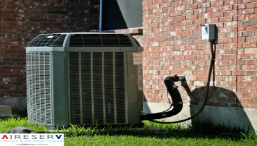 An outdoor air conditioning condenser unit installed beside a brick wall, with visible refrigerant lines and an electrical connection box.