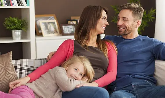 Couple smiling and staring at each other beside young daughter on couch in cozy living room.
