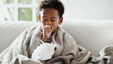 Child sitting on sofa coughing with tissues.