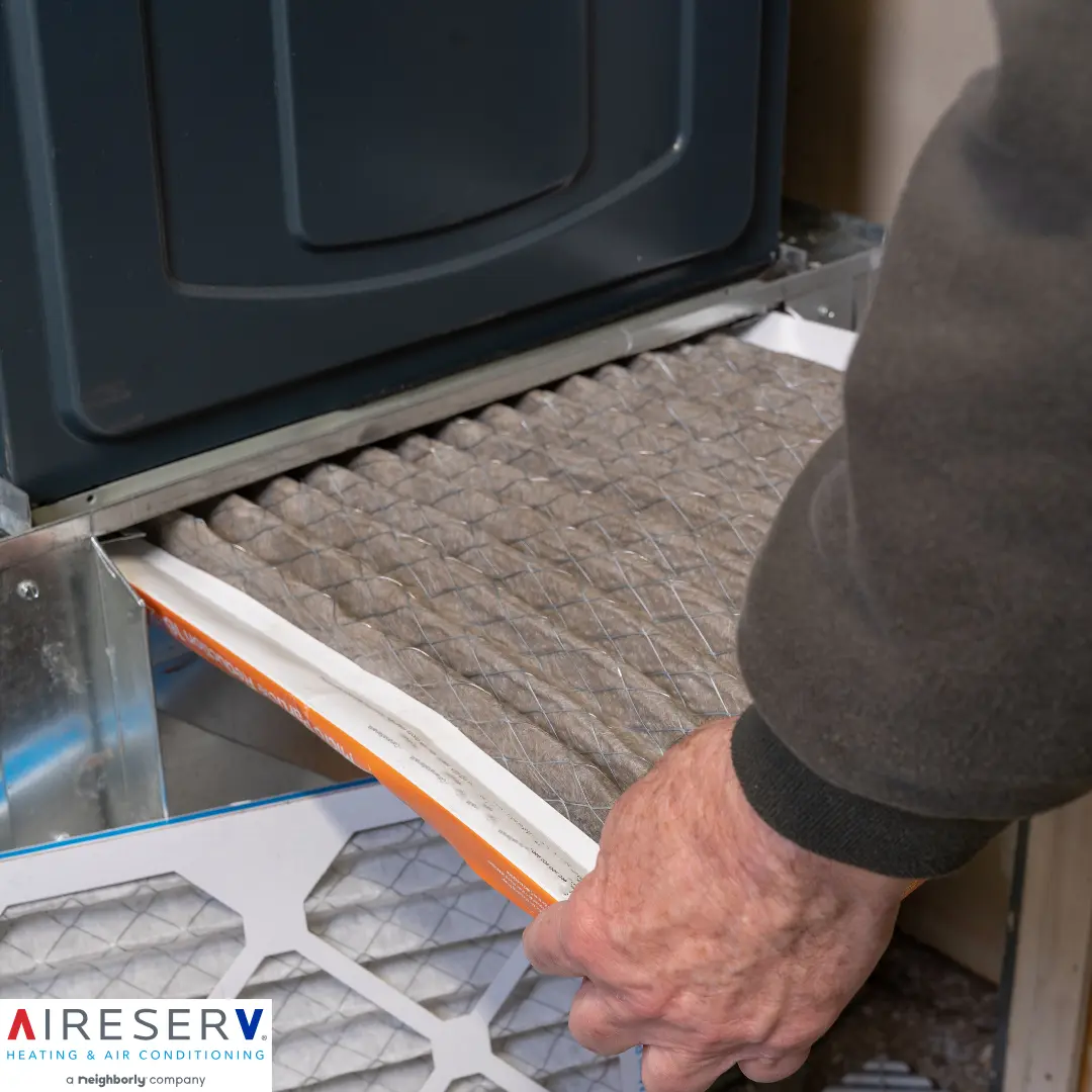 A hand removes a dirty, dust-covered HVAC filter from a furnace as a clean replacement filter sits in the foreground.