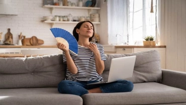 Woman suffers from an inoperative air conditioner, waving blue fan sitting on couch.