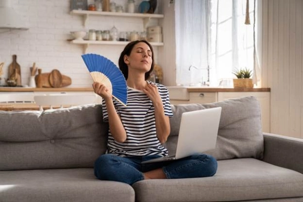 woman suffers from an inoperative air conditioner, waving blue fan sitting on couch.