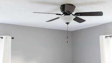 Black ceiling fan with light in brightly lit interior with gray walls and white curtains.