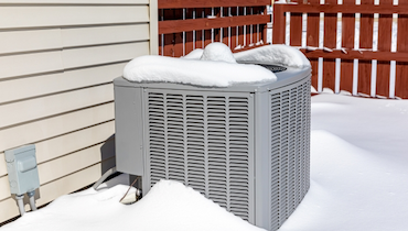 Central AC condenser unit covered in snow beside burgundy wood fence.