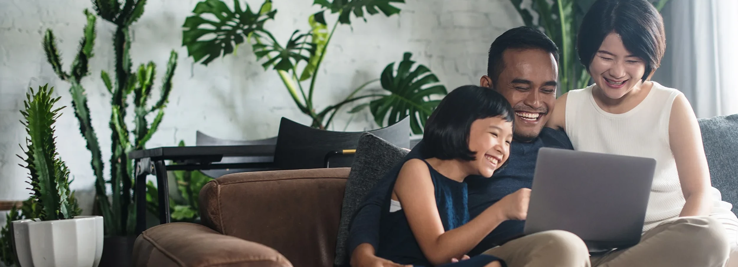 Smiling Asian couple and young daughter pointing and staring at laptop on couch in living room decorated with plants.
