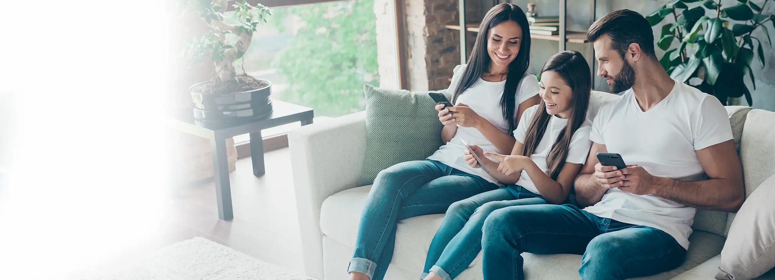 Smiling family sitting on couch, holding smart phones and viewing daughter's tablet.