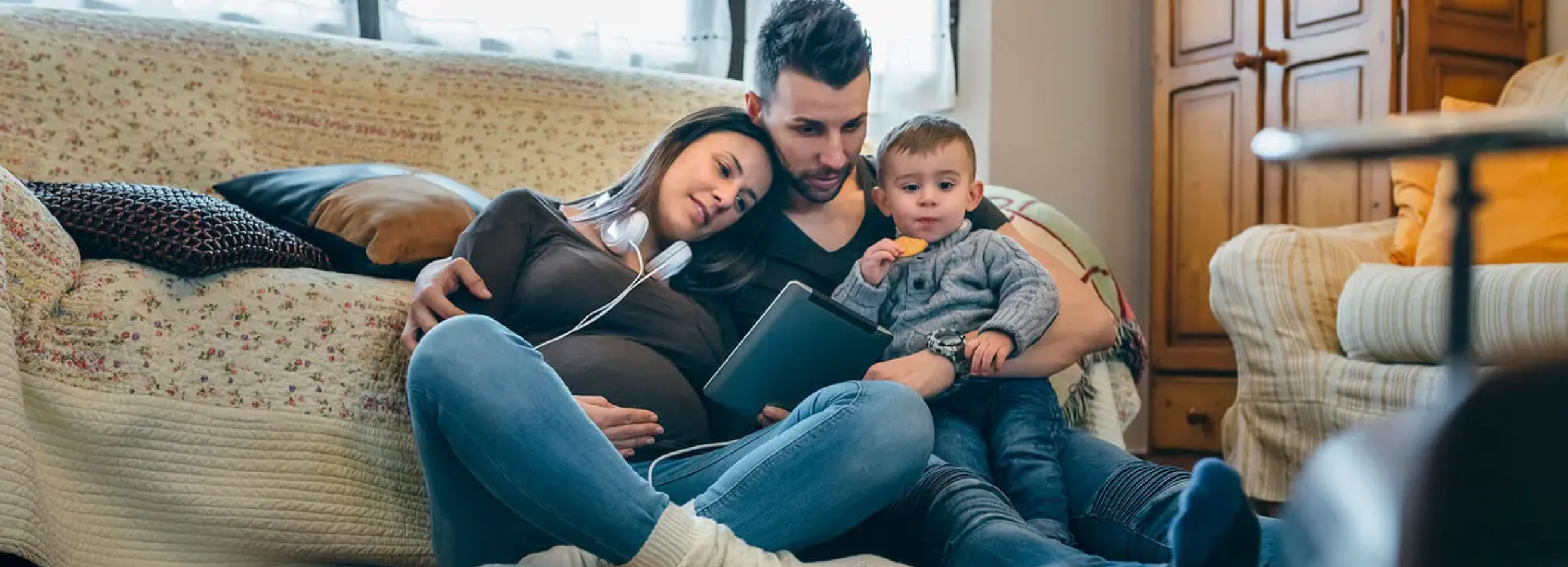 Pregnant mother sitting on living room floor holding tablet device beside husband and toddler.