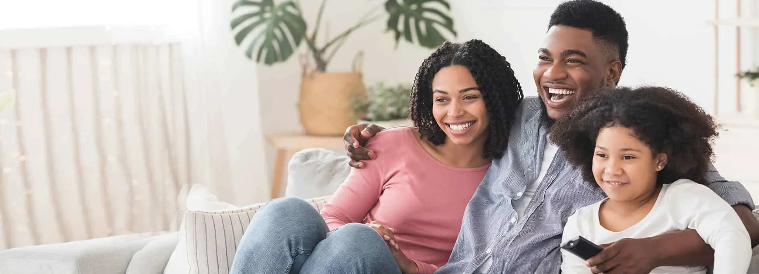 Young African American couple and daughter sitting on couch and smiling in brightly lit interior.