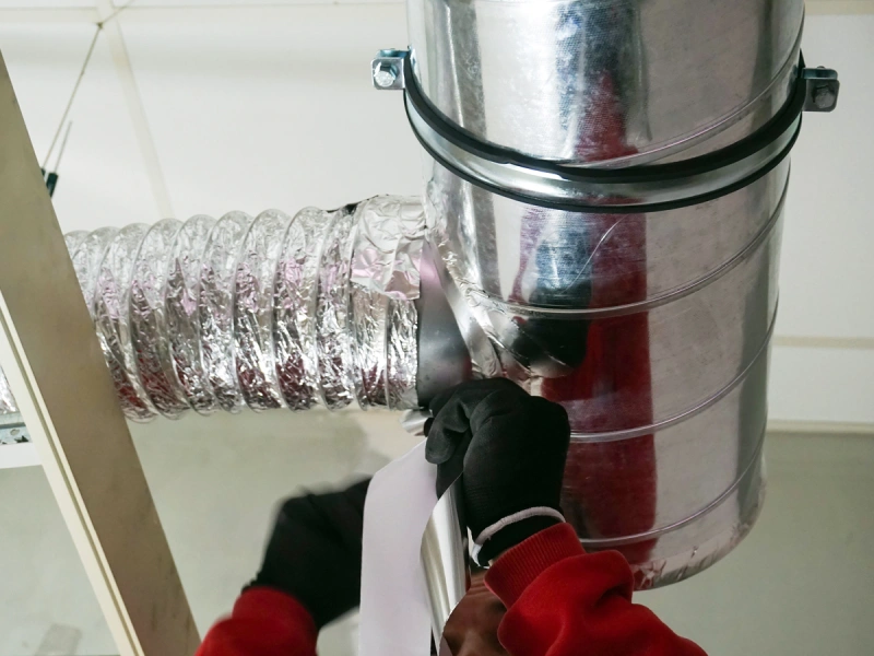 The gloved hands of an HVAC service professional in the process of using duct tape to secure a connection between a flexible duct and a rigid air duct.
