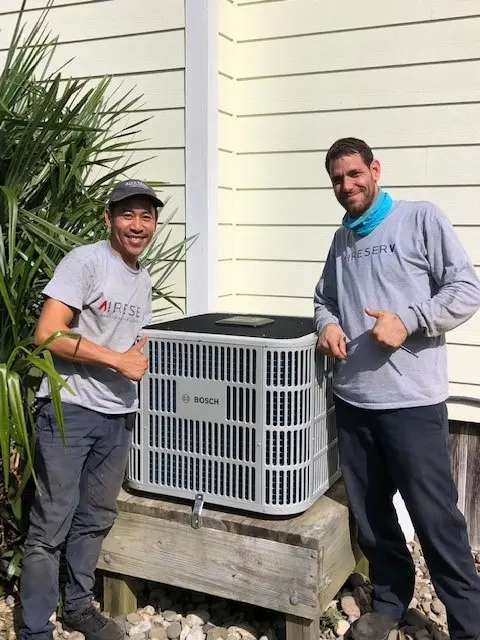Pair of male Aire Serv technicians giving thumbs up beside Bosch outdoor HVAC unit.