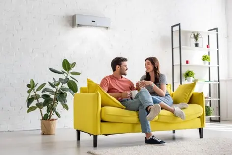 Man and woman sitting on yellow sofa with plants in background and mini-split AC unit installed on the wall behind them
