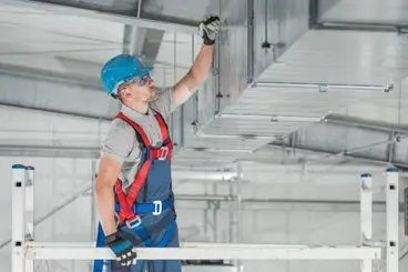 An HVAC technician stands on metal scaffolding and inspects metal ductwork on the ceiling of a commercial facility